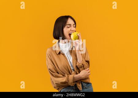 Concetto di stile di vita sano. Giovane donna che mangia gustosa mela su sfondo giallo, mordente frutta fresca deliziosa Foto Stock