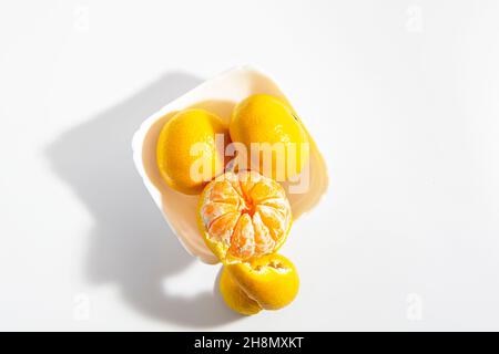 Due tangerini interi e una metà sbucciata in una ciotola bianca quadrata su sfondo bianco. Isolare. Primo piano. Foto orizzontale. Foto di alta qualità Foto Stock
