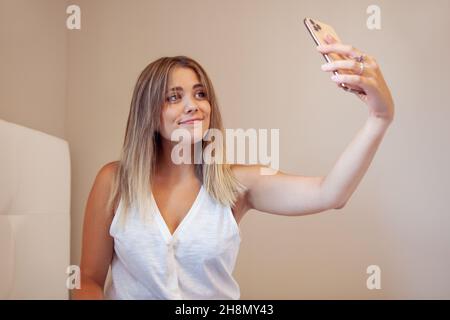Ragazza seduta sul letto guardando il mobile e sorridente. Donna che prende selfie e invia messaggi. Ragazza bionda. Foto Stock