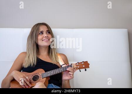 Donna che gioca l'ukulele seduto sul letto nella sua stanza a casa. Foto Stock