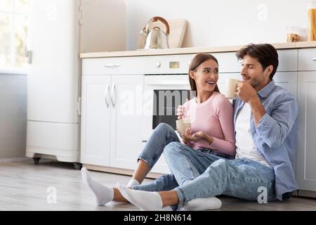 Soddisfatto felice giovane donna e ragazzo europeo con tazze di bevanda preferita si siede sul pavimento, parlare in cucina Foto Stock