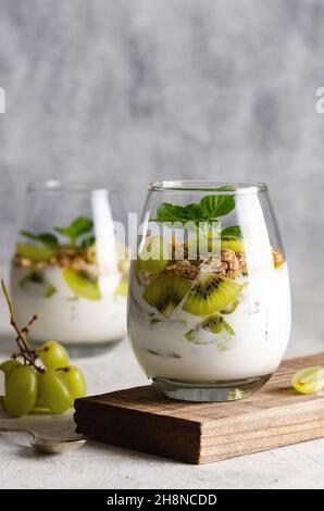 Due bicchieri di yogurt semplice con pezzi di kiwi freschi, uva bianca, muesli e foglie di menta su un vecchio pezzo di legno con fondo grigio. Foto Stock