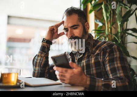 L'uomo adulto è seduto in un caffè e fa il suo programma quotidiano. Utilizza smartphone e appunti. Foto Stock