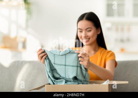 Acquisto di vestiti via internet. Felice giovane donna asiatica sboxing cartone di consegna pacchetto dopo lo shopping online Foto Stock