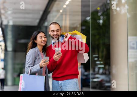 Giovane coppia interrazziale con smartphone in piedi su strada con borse shopping, acquistare merci online vicino al grande centro commerciale della città Foto Stock