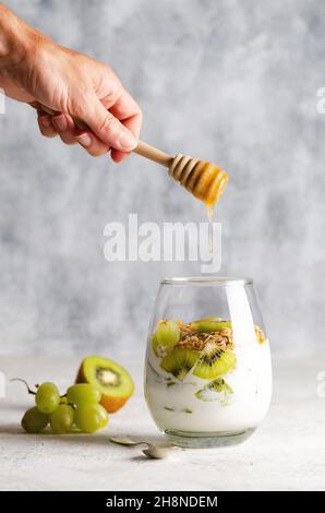 Yogurt bianco con pezzetti di kiwi freschi, uva bianca, granola, foglie di menta in un bicchiere, e un miele che sgocciola a mano con un contagocce al miele. Foto Stock