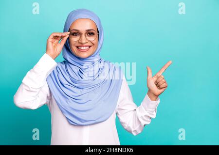 Foto di premuroso sicuro giovane donna indossare abiti arabo Headscarf braccio occhiali puntando spazio vuoto sorridente isolato di colore turchese sfondo Foto Stock