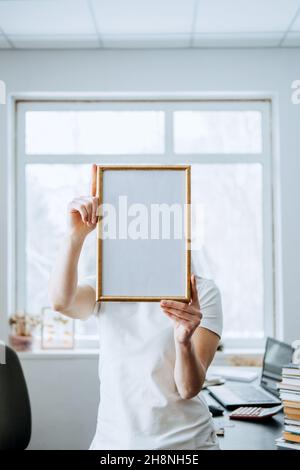 Cornice, poster, diploma, mockup certificato in mani femminili. Donna senza volto che tiene vuota cornice fotografica di legno bianco sul tavolo con il laptop e. Foto Stock