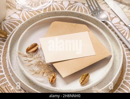 Tavolo da nozze con carta bianca e busta su placemat bohémien. Boho mockup carta da matrimonio. Sagoma beige tropicale con piante secche, luogo per Foto Stock