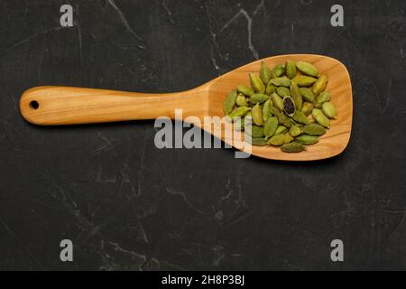 Cucchiaio di legno pieno di piccanti pod di cardamomo indiano, su uno sfondo di cemento scuro. Giacitura piatta Foto Stock