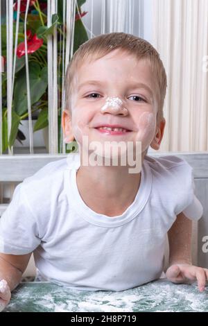 Un ragazzino emozionale carino, sporco di farina, ti guarda e ride. Gioca con la farina. Il bambino aiuta la madre con biscotti da forno a casa. Foto Stock