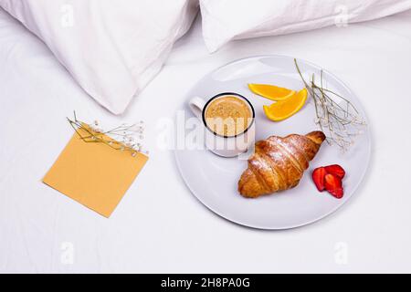 Piatto in ceramica con croissant, caffè, carta vecchia e ramo di fiori secchi sul letto. Colazione romantica a letto. Foto Stock