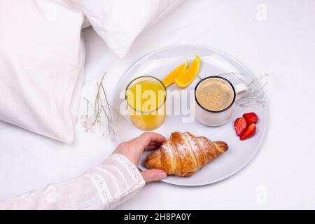 la mano della donna prende un croissant, una romantica colazione a sorpresa nel letto. Croissant, tazza di caffè, succo d'arancia sul piatto di ceramica. Dolce mattina. fl Foto Stock