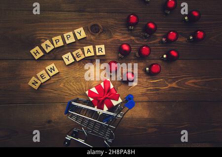Regalo in un piccolo carrello, iscrizione in lettere di legno felice anno nuovo e polpette di Natale multicolore su sfondo di legno. Sfondo di Natale scuro Foto Stock