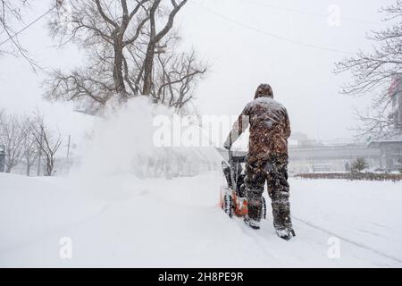 Un uomo in camouflage cammina sulla neve e sgomenta la neve con un spazzaneve a benzina. Vista posteriore. Foto Stock