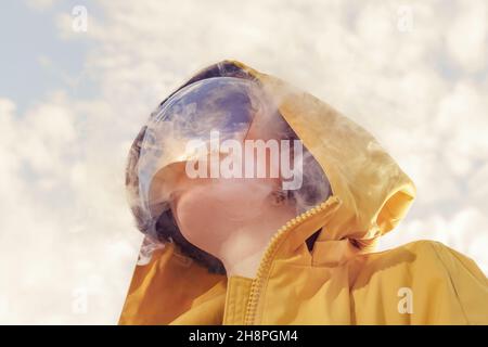 Una ragazza in un impermeabile giallo, una maschera a specchio che copre il suo volto e un riflesso del cielo in una maschera. Ritratto surreale futuristico Foto Stock