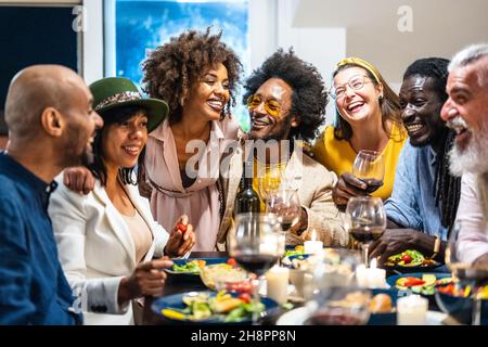 Persone diverse di età ed etnia che mangiano una cena vegana. Gruppo multietnico di amici divertirsi mentre si condivide un pasto in un caldo e accogliente hous Foto Stock