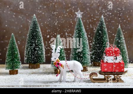 Slitta con regali di Natale e renna bianca accanto agli alberi di abete mentre sta nevicando su un tavolo di legno. Concetto minimo di Capodanno. Foto Stock