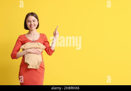 Giovane donna incinta con il bodysuit del bambino che punta a qualcosa su sfondo giallo Foto Stock
