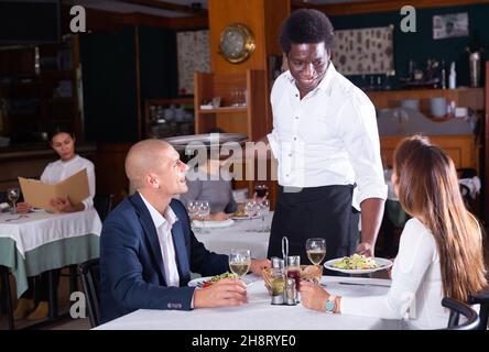 sorridente cameriere maschile portare ordine a uomo adulto e donna in restaurante Foto Stock