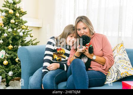 Una madre e sua figlia sono seduti su un divano nel soggiorno accanto all'albero di Natale che gioca con un piccolo cucciolo Foto Stock