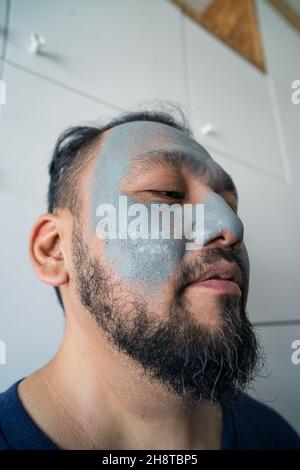 Ritratto di bellezza di un uomo asiatico maturato eccitato in piedi isolato su sfondo bianco, indossando facciale maschera di argilla cosmetica. Foto Stock