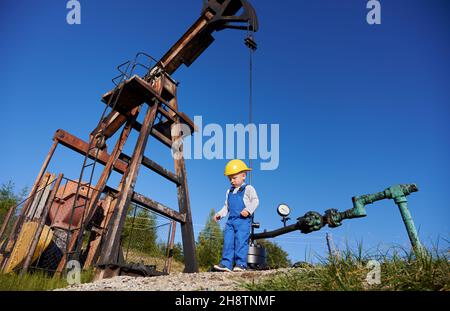 Vista dal basso di un piccolo ingegnere giovane vicino al carro di perforazione per olio. Ragazzo in tute contro il cielo blu, la pipeline di petrolio e il martinetto per pompa per l'estrazione di carburante naturale. Foto Stock