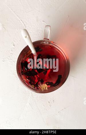 Una tazza di vetro con un cucchiaino di colore bianco con foglie di ibisco preparate su sfondo bianco. Tè rosso. Vista dall'alto. Rosella o fiori di rosa sudanese sono Foto Stock