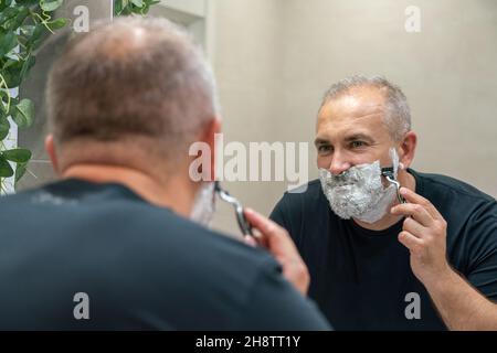 Uomo maturo dai capelli grigi che restyling la sua barba in casa utilizzando il rasoio Foto Stock