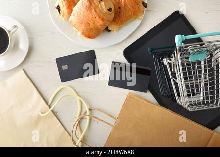 Concetto di shopping online con carrello, carte di credito e dispositivi su tavolo bianco in legno con colazione e shopping bag. Vista dall'alto. Foto Stock