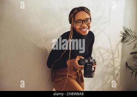 Giovane fotografo artistico che sorride alla macchina fotografica mentre si alza contro una parete nel suo ufficio. Felice giovane donna che tiene in mano una fotocamera dslr. C Foto Stock