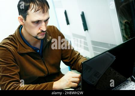 Ritratto di un programmatore al lavoro. Una scena autentica. Uno specialista IT lavora su un notebook nella sala server da 30-40 anni. Esecuzione della diagnostica o esecuzione di interventi di manutenzione. Sfondo. Foto Stock