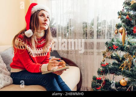 Giovane donna che scrive obiettivi elenco per il 2022 nuovo anno in taccuino a casa seduta da albero di Natale che indossa il cappello di Santa e caldo maglione. Foto Stock