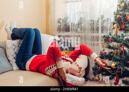 Annoiato deluso giovane donna sdraiata capovolta sul divano da albero di Natale che indossa Santa Hat sensazione triste, sconvolto, solitario. Da solo a casa per Capodanno Foto Stock