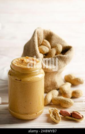 Vista a basso angolo su vaso in muratura di vetro con burro di arachide su tavolo di legno bianco con sacco a tela sul retro. Concetto di alimentazione sana Foto Stock