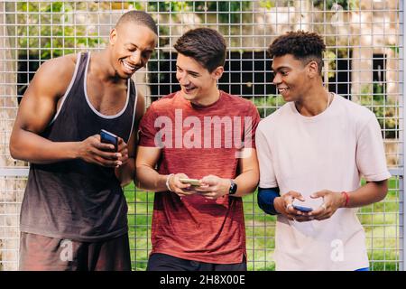 Allegri giovani atleti maschi multirazziali in smartphone sportivi e ridere mentre si trova vicino al cancello netto sullo stadio Foto Stock