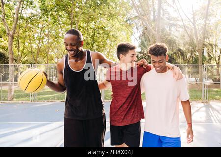 Allegro atleta maschile multirazziale in abbigliamento sportivo con palla di basket gialla ridendo a parlare e abbracciare mentre in piedi vicino alla recinzione nello stadio Foto Stock