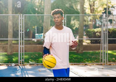 Sportivo afro-americano concentrato in abbigliamento sportivo in piedi sullo stadio sportivo e in possesso di palla di basket Foto Stock