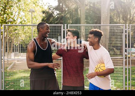 Allegro atleta maschile multirazziale in abbigliamento sportivo con palla di basket gialla ridendo a parlare e abbracciare mentre in piedi vicino alla recinzione nello stadio Foto Stock