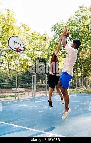 Amici etnici afroamericani giocatori correre insieme con la palla mentre giocano a basket sul campo sportivo in giornata di sole Foto Stock