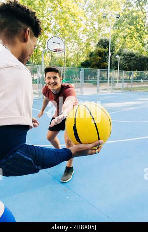 Amici multirazziali giocatori che corrono insieme con la palla mentre giocano a basket in campo sportivo in giornata di sole Foto Stock