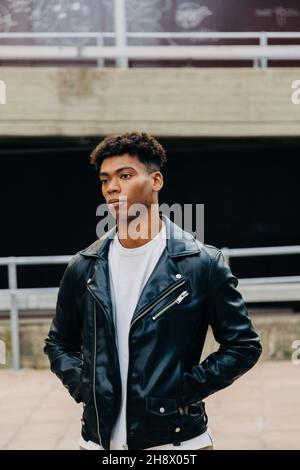 Giovane uomo nero pensieroso in giacca di pelle in piedi sulla strada con le mani in tasche contro ringhiera e guardare lontano Foto Stock