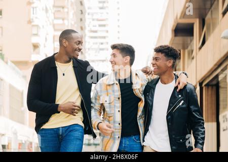 Giovani amici multirazziali sorridenti e si guardano mentre camminano vicino a edifici in strada Foto Stock