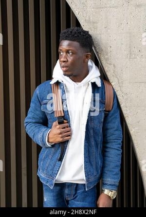 Maschio africano americano pensivo con capelli ricci scuri in abito denim in piedi vicino alla parete dell'edificio e guardando via Foto Stock