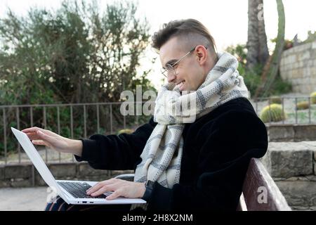 Allegro giovane maschio in abito alla moda con caldo sciarpa e occhiali sorridenti e con videochiamata via computer portatile seduto su panchina nel parco tropicale Foto Stock