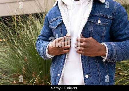 Maschio africano americano tagliato e irriconoscibile in giacca e felpa in denim in piedi vicino al verde cespuglio in strada della città di giorno Foto Stock