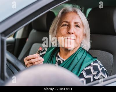 Donna anziana in abbigliamento elegante applicando il rossetto mentre si siede sul sedile del conducente di un'auto moderna Foto Stock