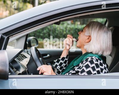 Vista laterale anziani Donna in abbigliamento elegante applicazione rossetto guardando nello specchio retrovisore mentre si siede sul sedile del conducente di un'auto moderna Foto Stock