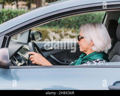 Ragazza anziana allegra in elegante vestito guida moderna macchina lucida in giorno di sole e guardando via Foto Stock