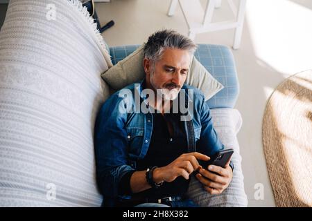 Dall'alto di elegante anziano maschio in abiti denim poggiato su un comodo divano in soggiorno di appartamento loft e utilizzando smartphone Foto Stock